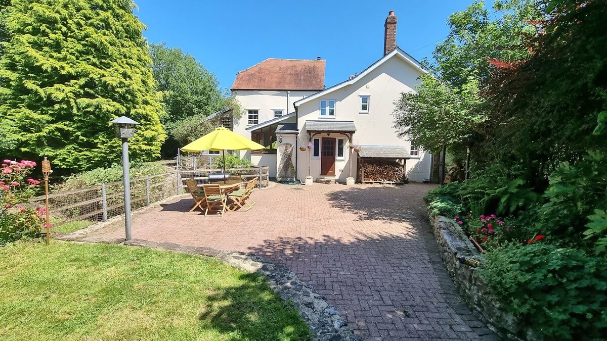 A spacious outdoor patio area is depicted, featuring a large yellow umbrella providing shade over a wooden dining table and chairs. The surrounding garden is filled with greenery, and a stone wall enhances the landscape. A woodpile is visible beside the entrance of the house.