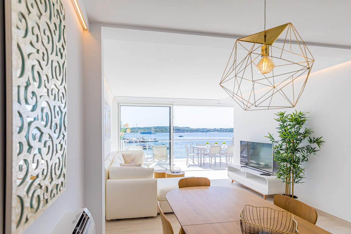 A modern living area features large glass sliding doors that offer expansive views of the bay. Light-colored furniture complements the bright space, while a dining table is set for gatherings. An accent wall with intricate design adds visual interest, and greenery is present from a nearby plant.