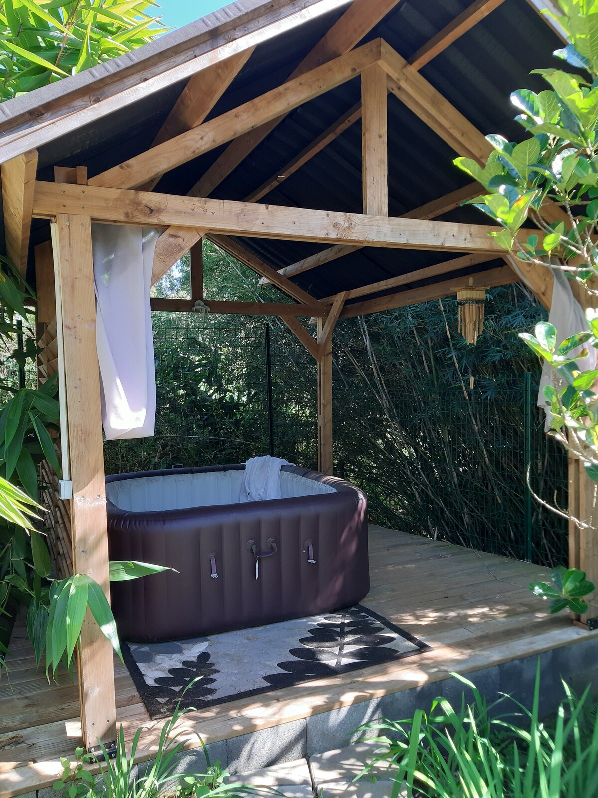A shaded wooden gazebo is surrounded by lush greenery, providing a secluded space. Within the gazebo, a hot tub is placed on a wooden deck. A soft towel is neatly draped over the edge of the tub, inviting relaxation. Natural light filters through the surrounding plants.