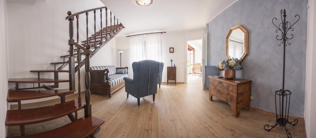 A spacious living area features a wooden staircase leading to the upper level. A blue armchair is positioned across from a striped sofa, complemented by natural light filtering through large windows. A wooden side table and a decorative mirror add to the room's elegance.