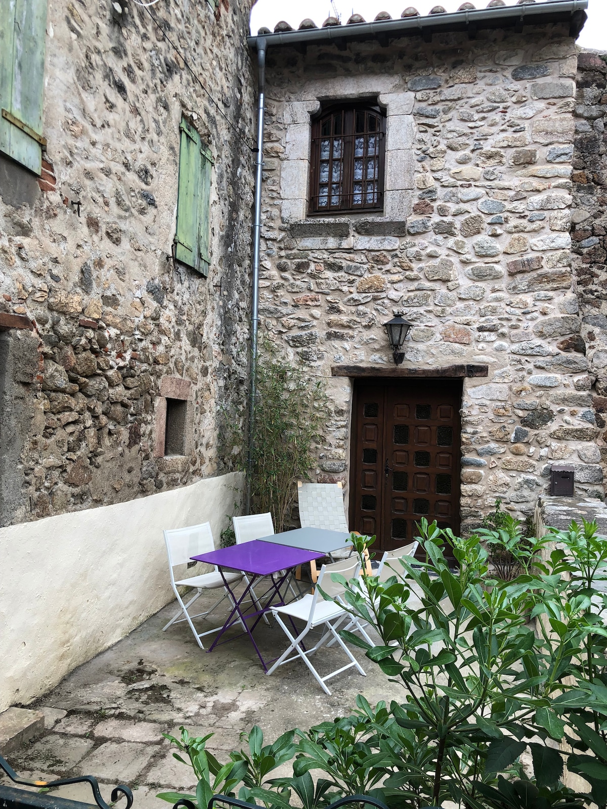 A small outdoor space is featured, showcasing a purple table accompanied by four chairs. The backdrop consists of a rustic stone wall with green shutters and a wooden door, surrounded by lush greenery. Natural stone textures create a historically rich atmosphere.
