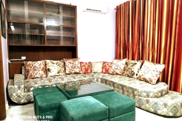 A spacious lounge area is furnished with a large, U-shaped sofa featuring decorative cushions in various patterns. A glass-top coffee table occupies the center of the seating arrangement. Rich, warm curtains frame the window, enhancing the inviting ambiance.