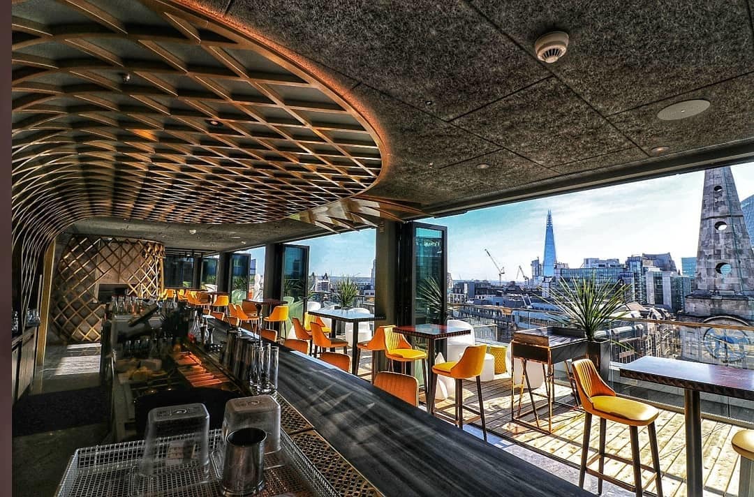 A rooftop bar features a modern design with a curved ceiling and large windows, offering expansive views of the city skyline. Stylish yellow chairs are arranged around tables, while the sleek bar area showcases various equipment and plants for added greenery.