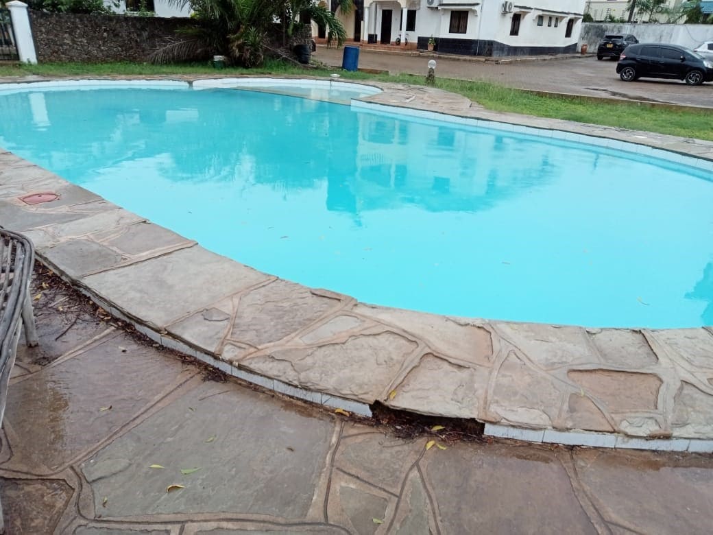 A refreshing swimming pool is framed by a stone patio, featuring clear turquoise water that invites relaxation. Surrounding foliage adds a natural touch, while the serene setting is enhanced by a glimpse of the apartment complex in the background.
