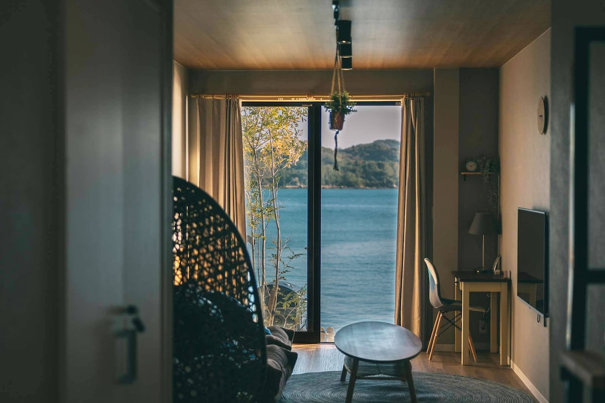 A spacious living area is visible, featuring a large glass window that offers a clear view of the ocean. Natural light fills the space, highlighting a circular wicker chair next to a coffee table. A small desk with a chair is positioned to one side, while greenery can be seen outside.