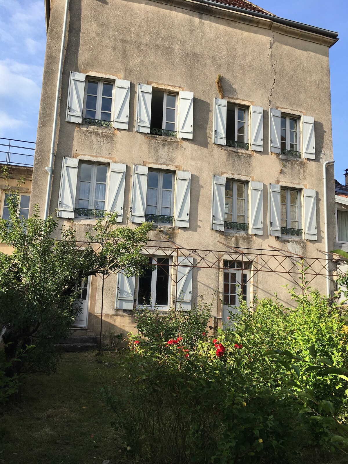 A historic three-story building is shown with multiple windows adorned with blue shutters. The facade features a mix of light and shadow, while a small garden area with greenery and blooming flowers occupies the foreground, inviting a sense of outdoor tranquility.
