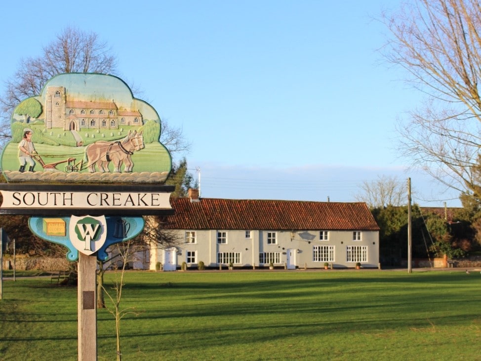 The image depicts a charming village sign for South Creake, showcasing a painted scene of a man with a horse-drawn cart. In the background, a picturesque building with large windows and a tiled roof is visible, set against a clear blue sky over a green field.