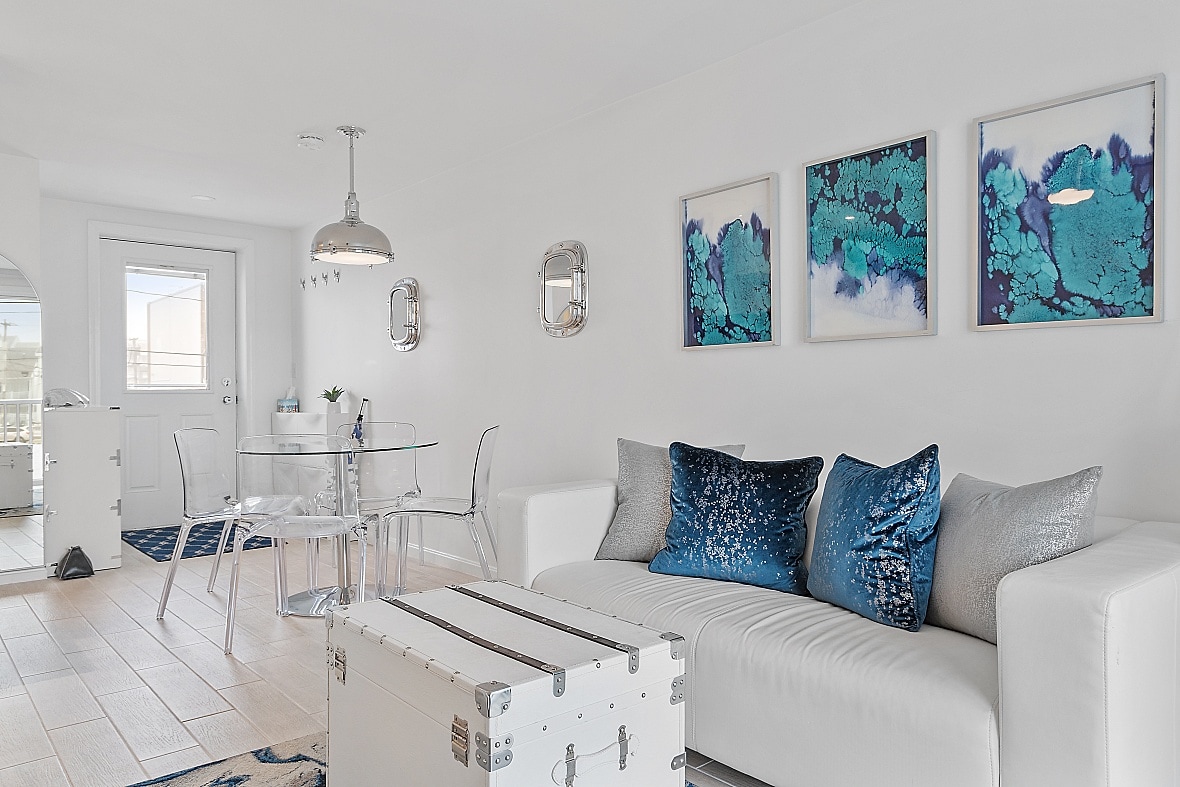 A bright, airy living area features a white sofa adorned with plush cushions in shades of silver and blue. A glass dining table accommodates four. Three vibrant wall art pieces in teal tones enhance the modern decor. A door leads to an outdoor space.