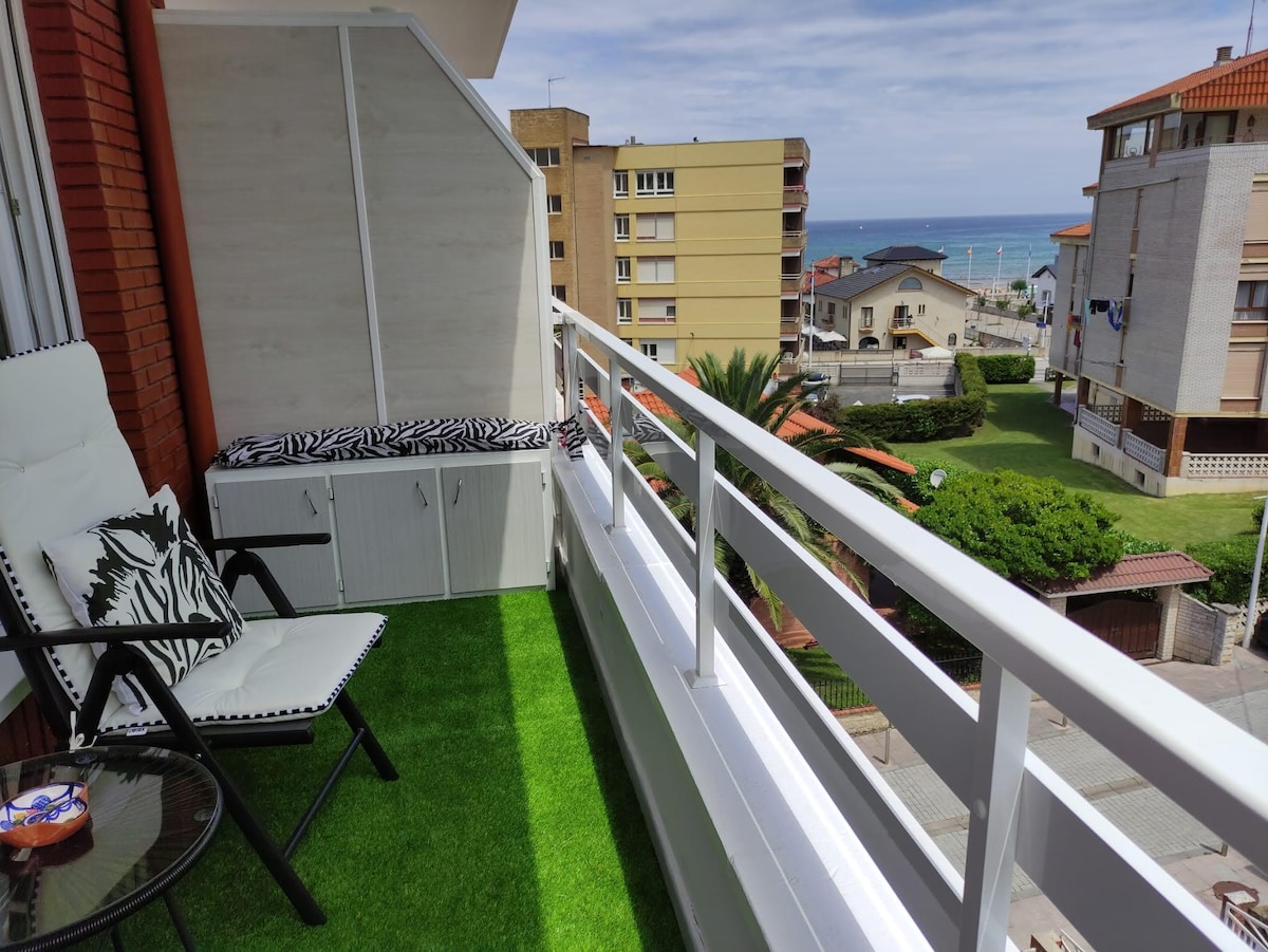 A terrace is presented, featuring a comfortable seating area with a black and white chair. A cushioned bench is positioned against the wall, with vibrant green turf underfoot. The view includes nearby buildings, palm trees, and the sea in the distance, enhancing the relaxing outdoor space.