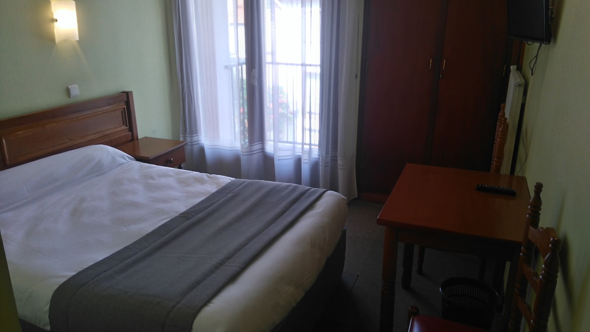 A comfortable sleeping area features a spacious bed with a light-colored blanket. Two windows with sheer curtains allow natural light to fill the room. A small wooden desk and chair are positioned nearby, complementing the wooden accents of the furniture.