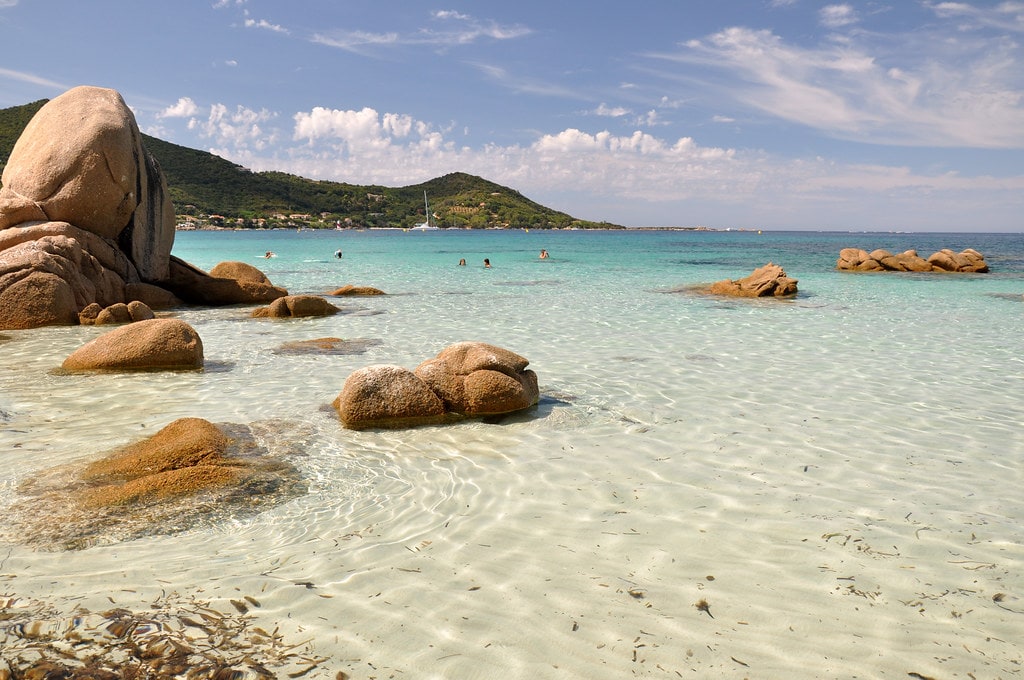 A serene beach scene is captured, featuring clear turquoise waters gently lapping against smooth, warm rocks. The shoreline is adorned with sandy patches interspersed with rocks. A distant view of lush hills and a few small boats reflects a tranquil atmosphere under a bright blue sky.