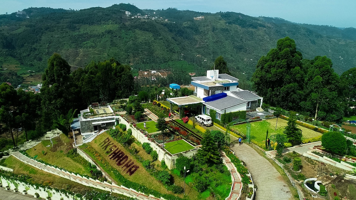 The aerial view reveals a spacious villa surrounded by lush greenery and meticulously landscaped gardens. Paths meander through various plant beds, leading towards a modern building featuring large windows. Hills in the background add to the serene setting, indicating a peaceful retreat.