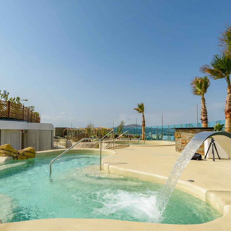 A recreational area features a curving water feature with gentle jets, surrounded by a spacious deck and several palm trees. Clear skies and distant views of the landscape enhance the relaxing atmosphere of this outdoor space.
