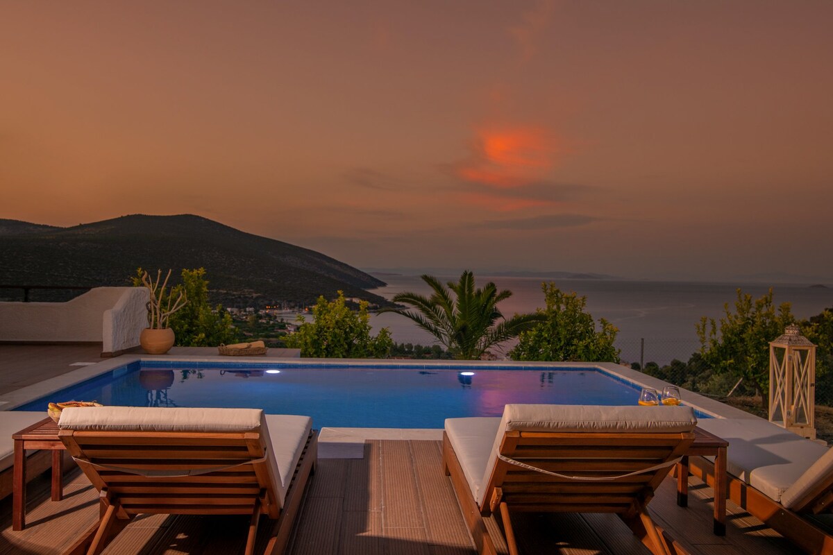 A serene outdoor space is showcased with two loungers beside a tranquil swimming pool. A panoramic view of the surrounding hills and waters is visible under a warm sunset sky. Soft lighting highlights the pool area, creating a welcoming atmosphere.