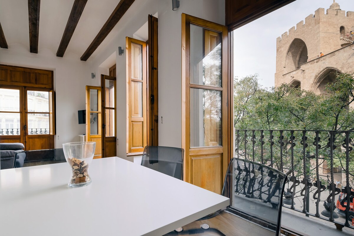 A dining area with a white table and transparent chairs is positioned near large windows that open to reveal views of the historic Torres de Serranos. Natural light fills the space, highlighting the wooden beams and elegant decor.