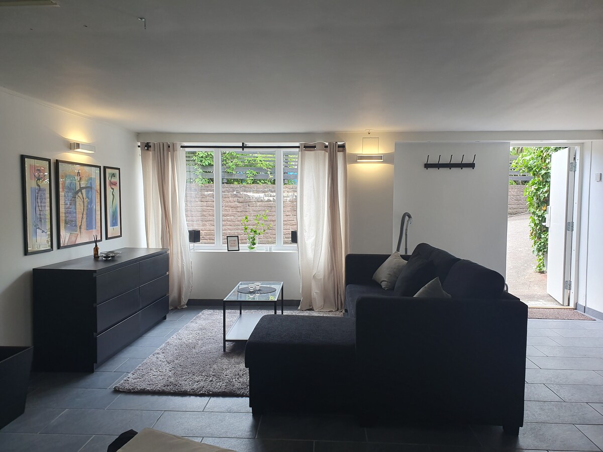 A spacious living area is highlighted by a comfortable dark sofa and a glass coffee table. Soft curtains frame the windows, allowing natural light to fill the room. Artwork adorns the walls, and an entryway is visible on the right.
