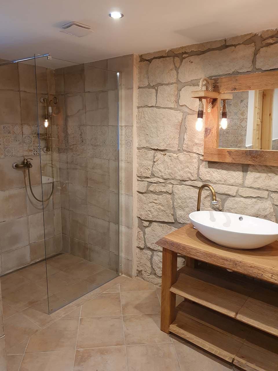 A modern bathroom features a glass-enclosed shower with a sleek bronze showerhead. A wooden vanity with a round basin is positioned under a wooden-framed mirror, complemented by industrial-style light fixtures. Exposed stone walls add character to the space.