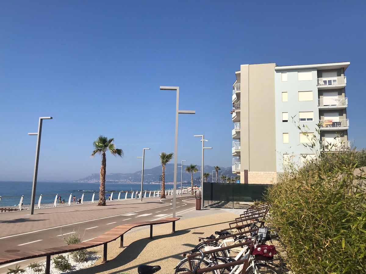 A beachfront view displays a modern building alongside a palm-lined walkway. Bicycles are parked in the foreground, while a clear blue sky complements the serene sea in the background. The landscape is designed for pedestrian access, enhancing the coastal atmosphere.