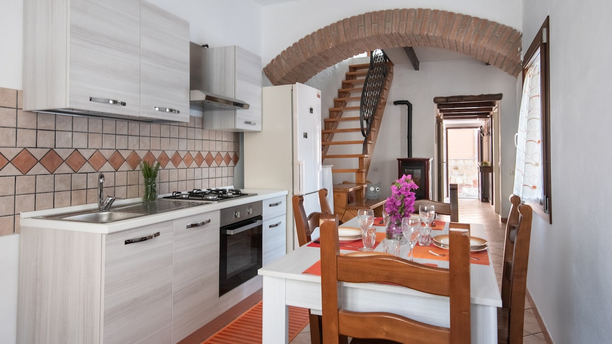 The kitchen area features modern cabinetry and a gas oven, paired with a spacious dining table set for four. A staircase with a decorative railing leads to the upper floor, while natural light illuminates the space through a nearby window. Vibrant flowers add a touch of color to the setting.