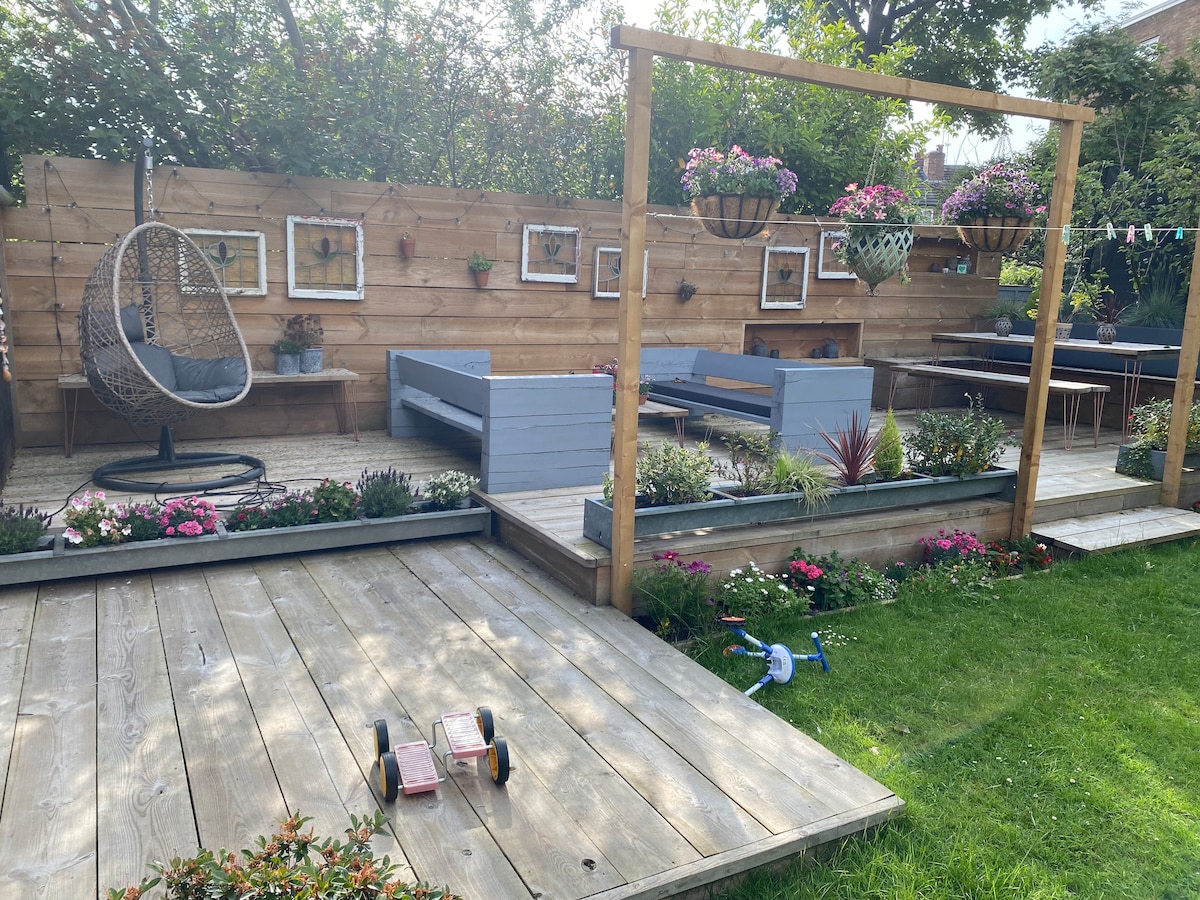 A spacious garden area features wooden decking and a range of seating options, including a hanging chair and a raised platform with benches. Vibrant flowers and greenery enhance the surroundings, while play equipment is visible on the grass, inviting relaxation and outdoor activities.
