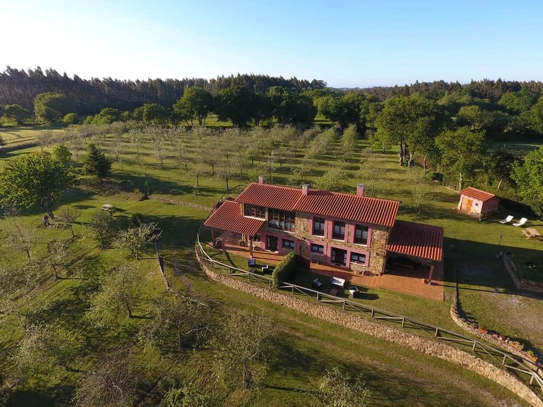 An aerial view captures a two-story house with a red tiled roof surrounded by a lush green landscape. Fruit trees are visible in neat rows, with open space highlighted by scattered vegetation. The property is enclosed by a rustic wooden fence, enhancing the natural setting.