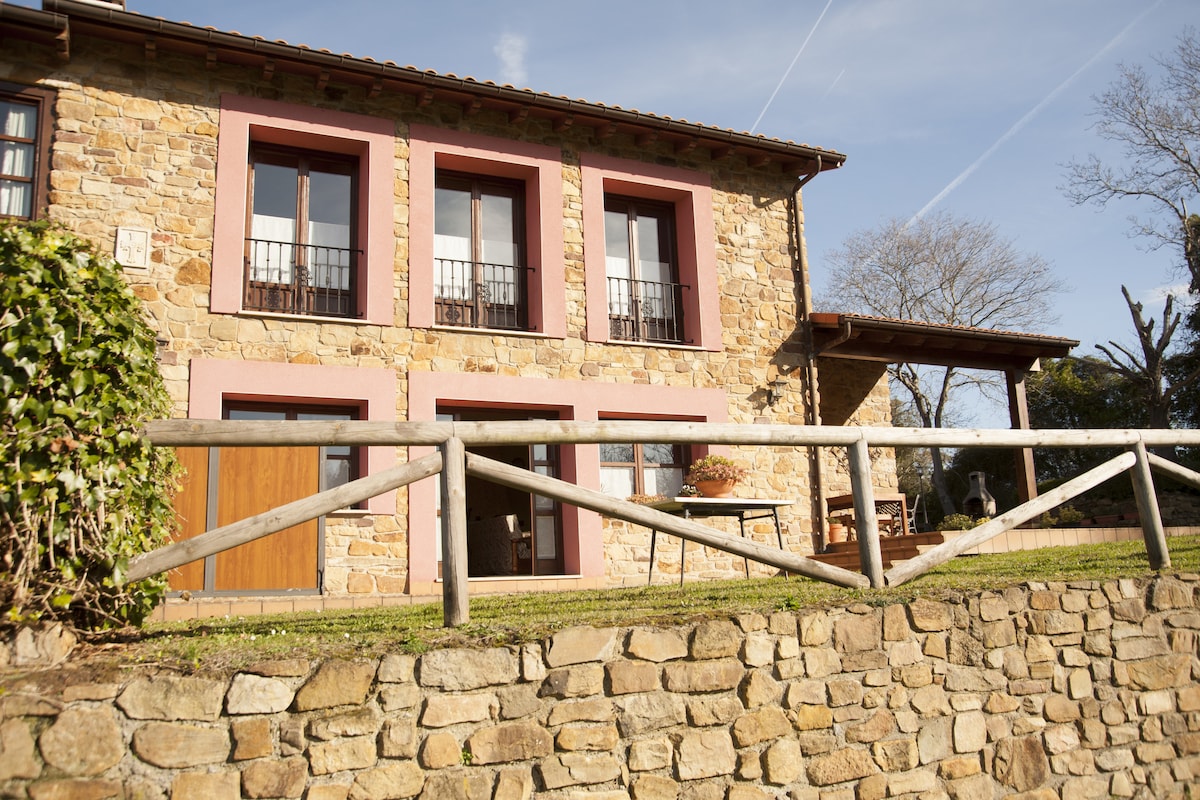 The exterior of a stone house is highlighted, displaying two upper windows with balconies and a lower wooden door. A railing made of wooden logs runs along the grassy area, which is bordered by a low stone wall. The surrounding landscape features trees under a clear sky.
