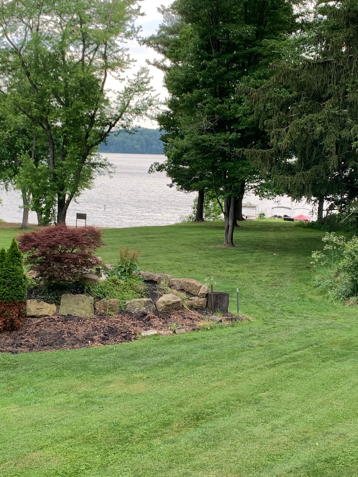 A serene lakeside view is presented, featuring a gently sloping grassy area leading to the water. Various trees provide shade, and a landscaped flower bed adds texture in the foreground, contrasting with the calm lake in the distance.