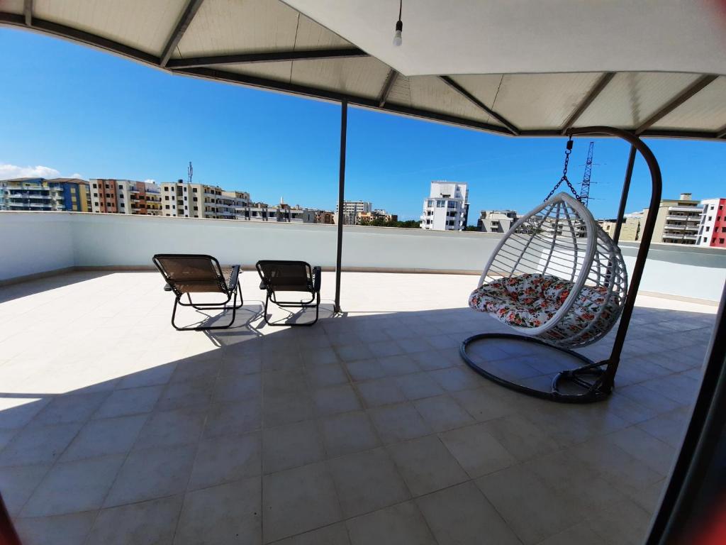 A spacious private terrace showcases two lounge chairs and a hanging swing chair with colorful cushions. The open area is illuminated by natural light, highlighting the surrounding cityscape under a clear blue sky.