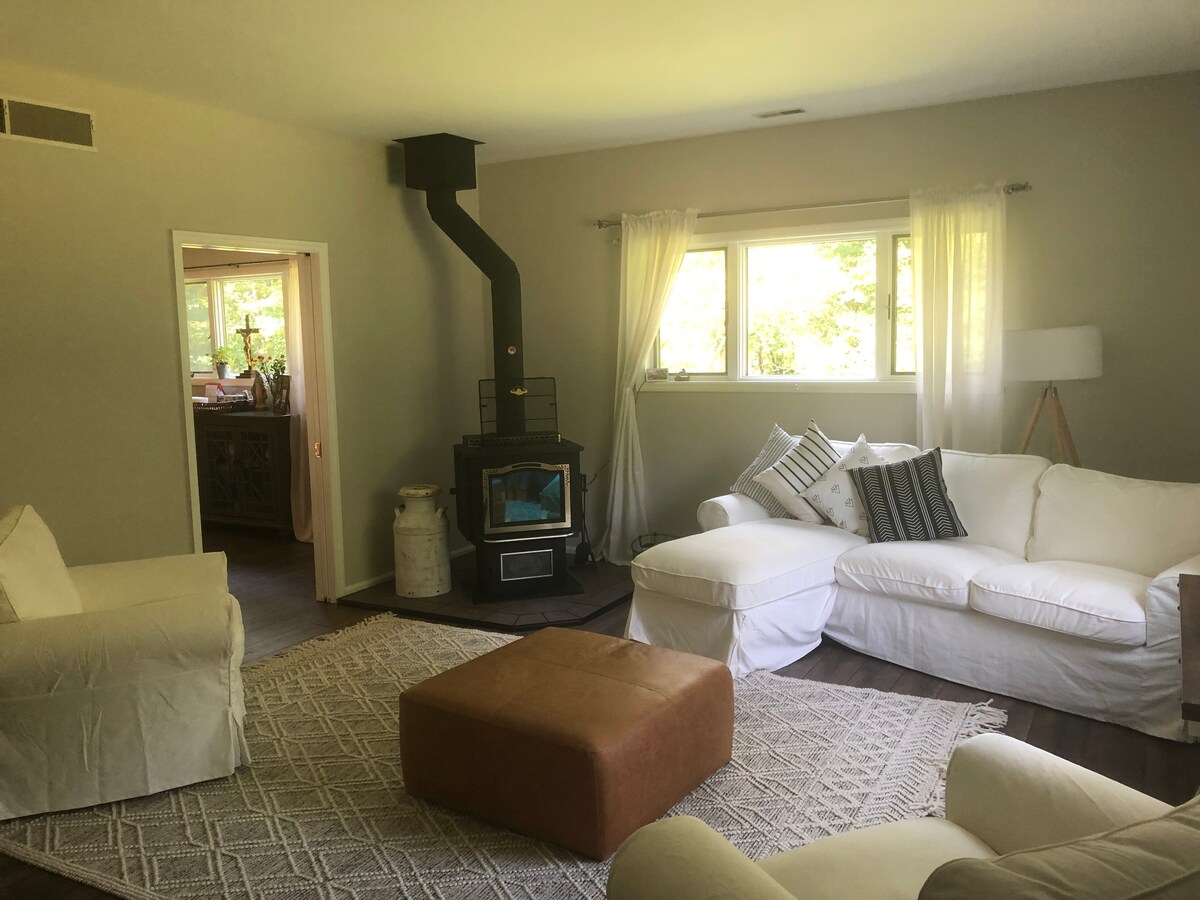 A bright living area is furnished with a white sectional couch and a comfortable armchair. A soft, textured rug covers the floor, and a wood-burning stove is positioned in the corner. Natural light filters through the windows, enhancing the cozy ambiance.