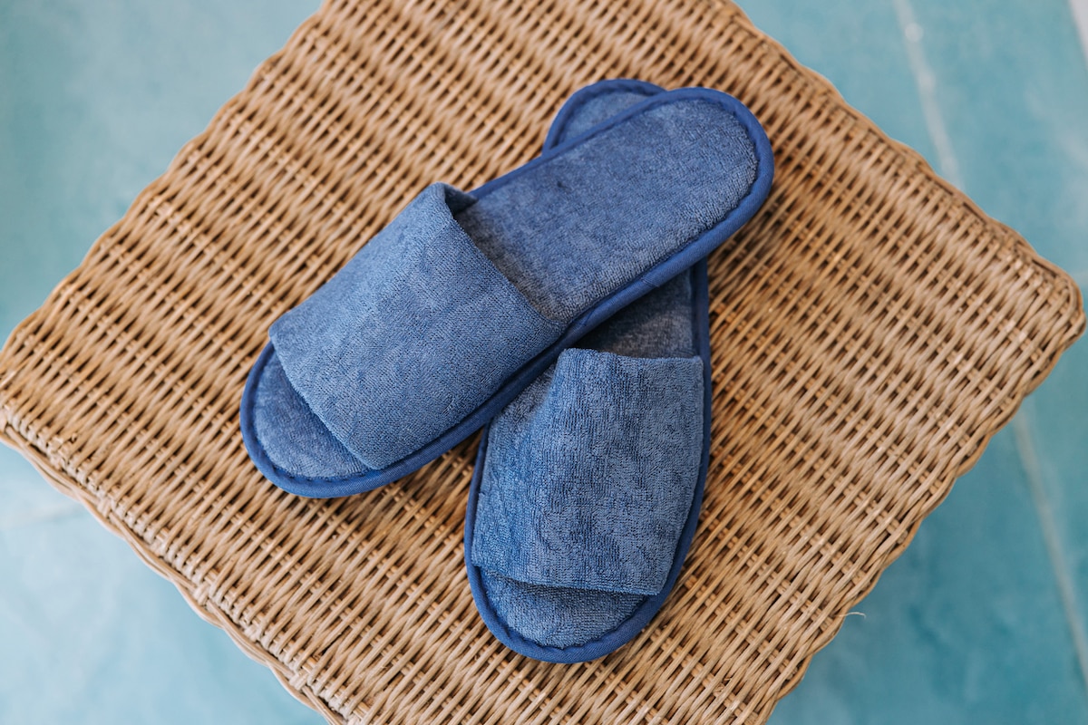 A pair of blue fabric slippers is placed on a woven wicker surface. The slippers feature a simple design with open toes and a wide, soft strap, ideal for comfort during stays.