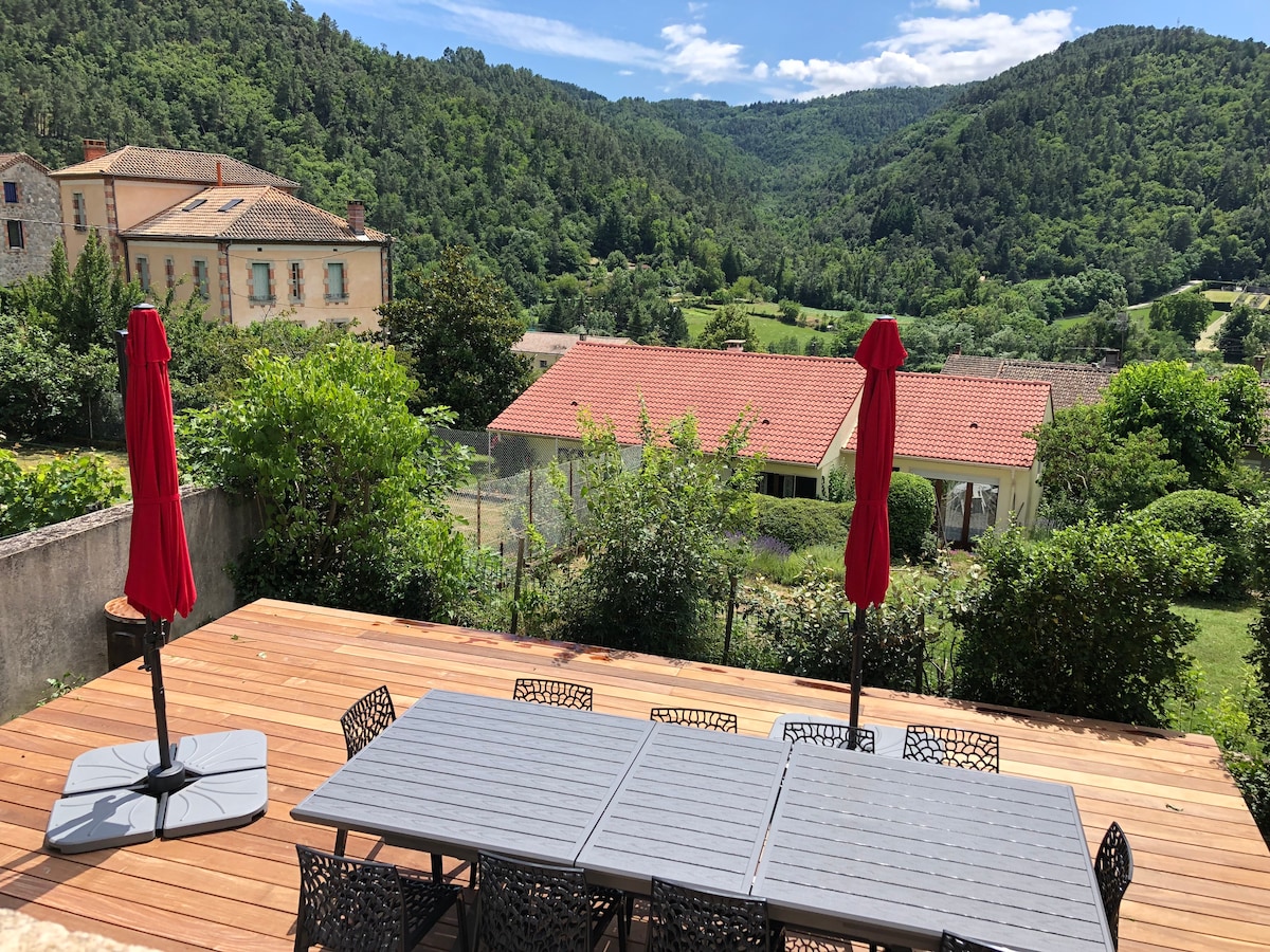 A large wooden terrace is shown, featuring a long dining table surrounded by several chairs. Two red umbrellas provide shade, and lush greenery is visible in the background, along with rolling hills and traditional buildings.