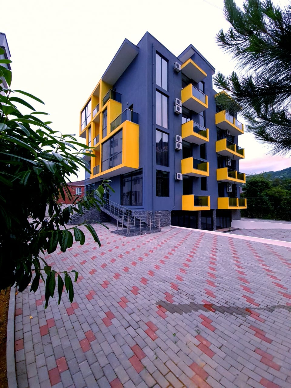 A modern three-story building features large glass windows and a striking yellow accent on the exterior. Multiple balconies are visible, showcasing an innovative design. Surrounding greenery and a paved pathway lead to the entrance, enhancing the contemporary look of the establishment.