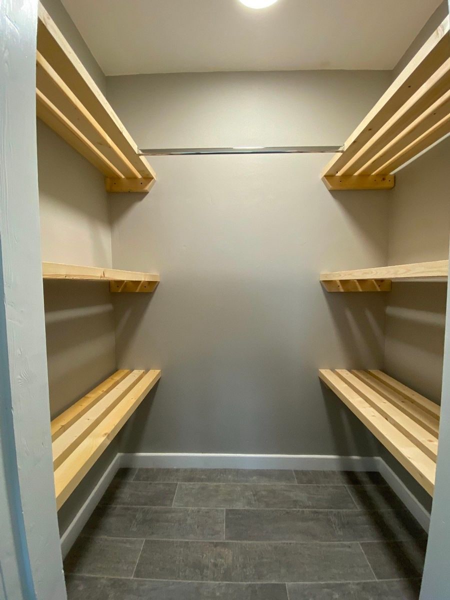 A spacious storage area features wooden shelving arranged on opposing walls. The light grey walls and tiled flooring contribute to a clean and organized appearance. Bright overhead lighting enhances visibility within the room.