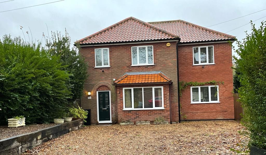 A two-story brick house features a tiled roof and several windows. Lush greenery surrounds the property, providing a sense of privacy. A gravel driveway leads to the entrance, enhancing the home's welcoming appearance within its serene setting.