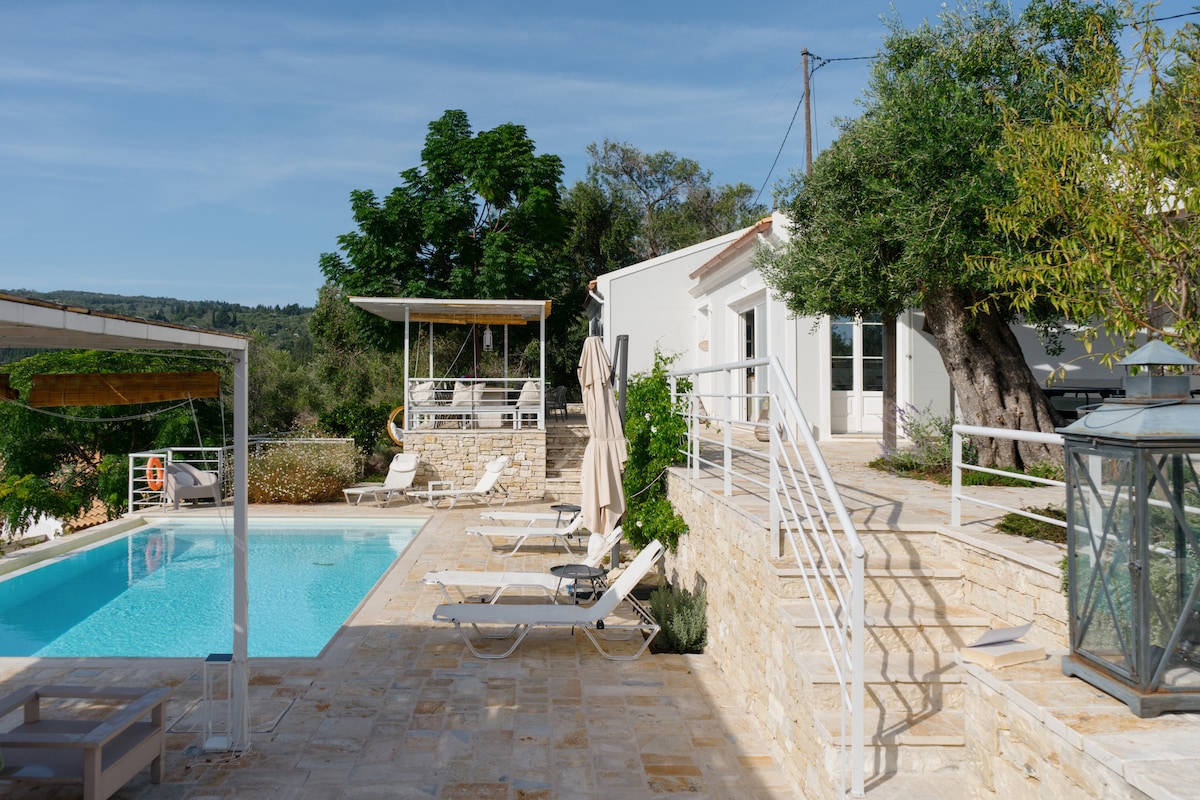 A spacious outdoor area features a private infinity pool surrounded by sun loungers. Stone pathways lead to terraced sections with greenery. The villa's exterior showcases large windows, providing views of the serene landscape. A shaded lounge area is also visible, inviting relaxation.