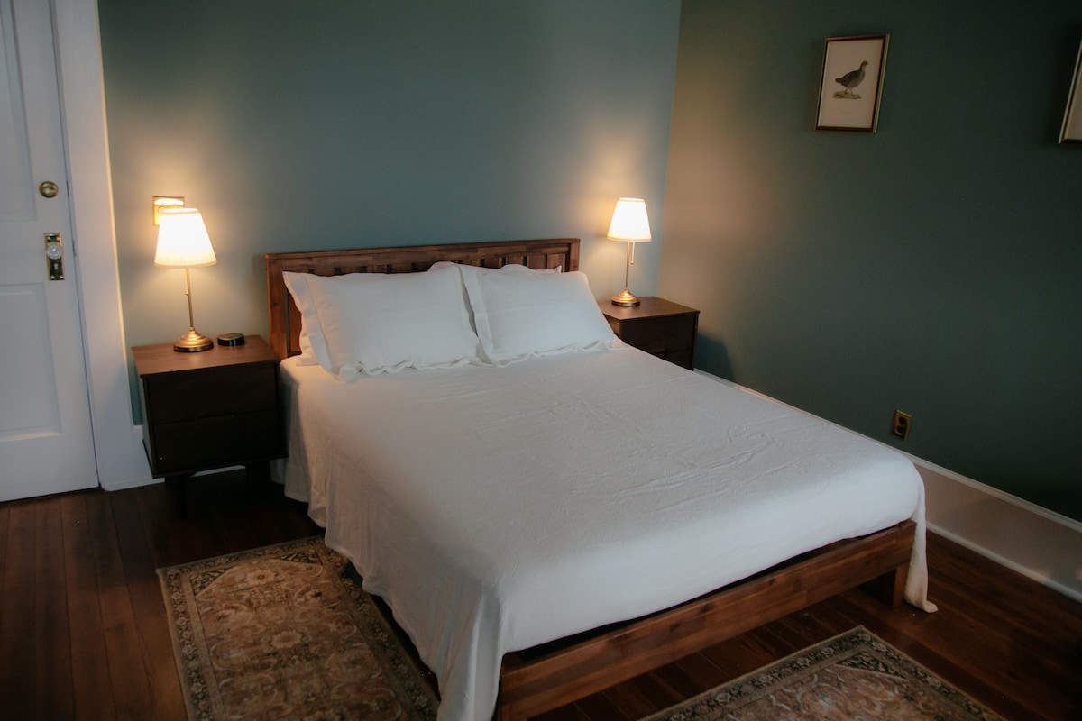 A serene bedroom is shown, featuring a spacious bed with white linens and two bedside lamps casting a warm glow. The walls are painted in a calming green hue, complemented by a decorative area rug beneath the bed.