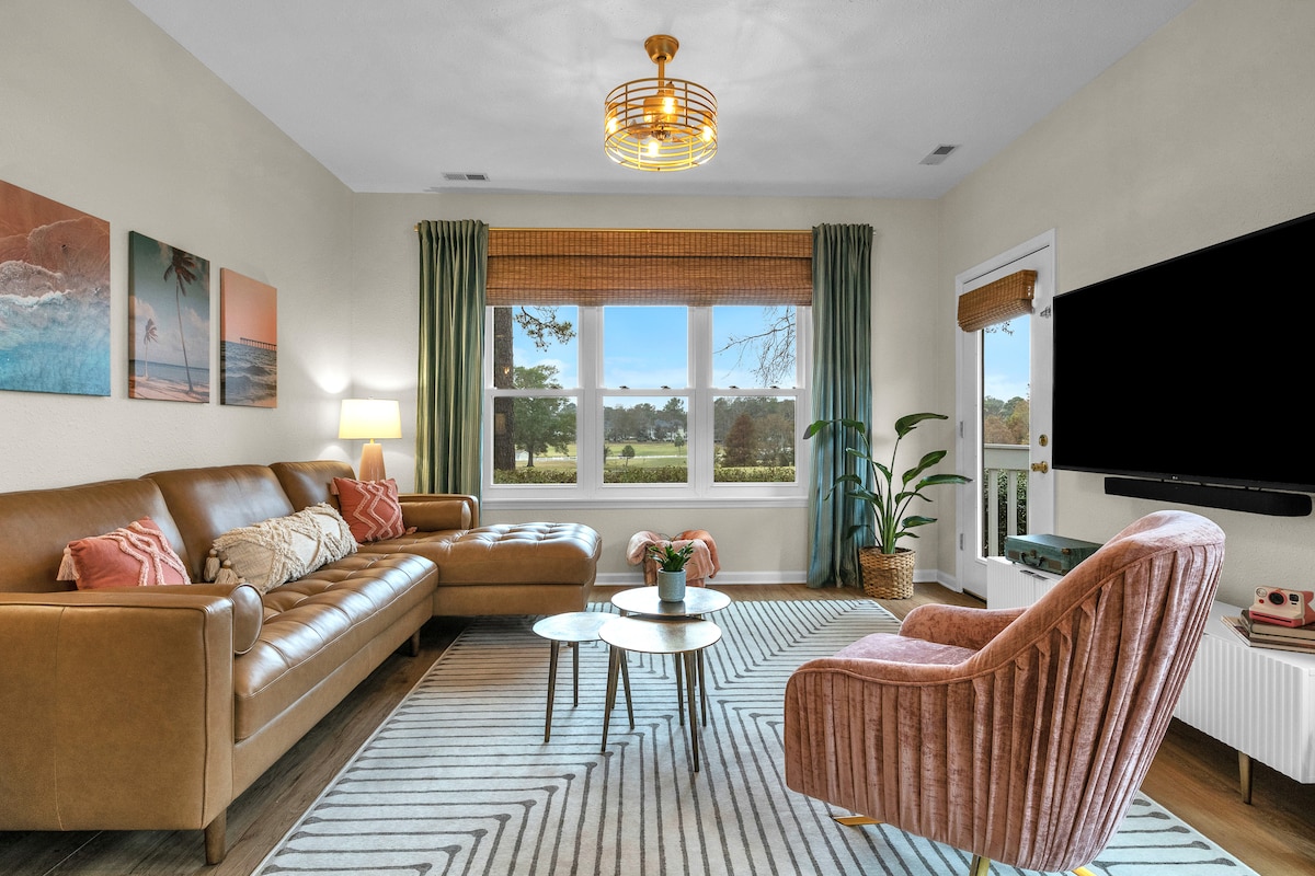 A comfortable living area features a brown leather sofa and a pink velvet chair, positioned around a circular coffee table. Light streams through large windows, revealing views of lush greenery. Art pieces adorn the walls, and plants add a touch of nature to the space.