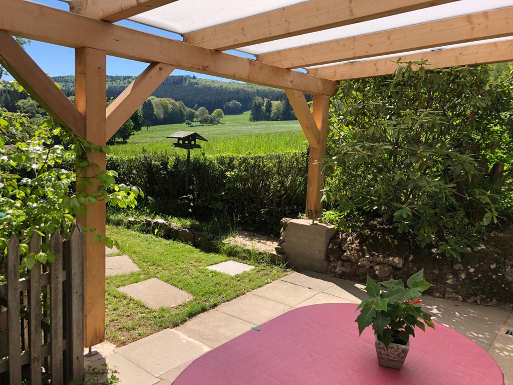 An outdoor area is covered by a wooden pergola, providing shade over a round table set on tiled flooring. Green fields and trees are visible in the background, while a bird feeder stands nearby amid lush greenery.
