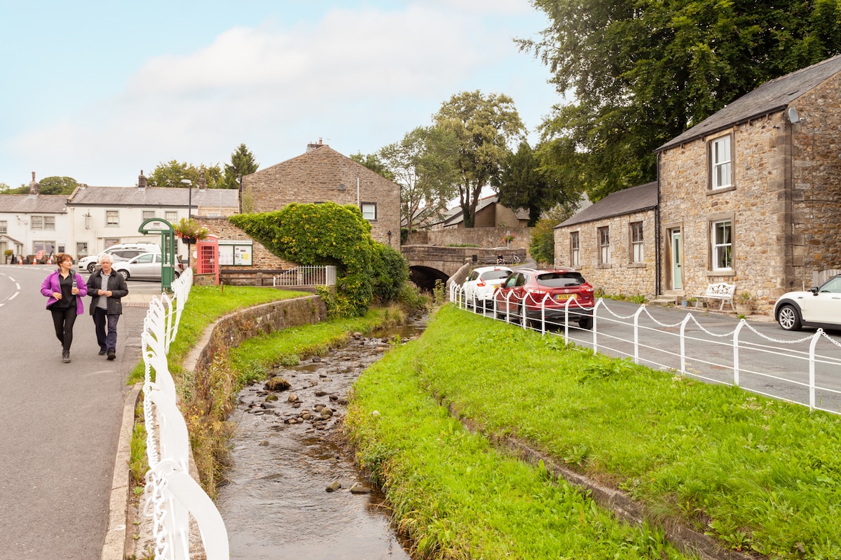 A scenic village view is displayed, featuring traditional stone buildings alongside a gentle stream. Walking paths are visible, with pedestrians enjoying the area. Vehicles are parked along the street, and lush greenery borders the flowing water, enhancing the charm of the surroundings.