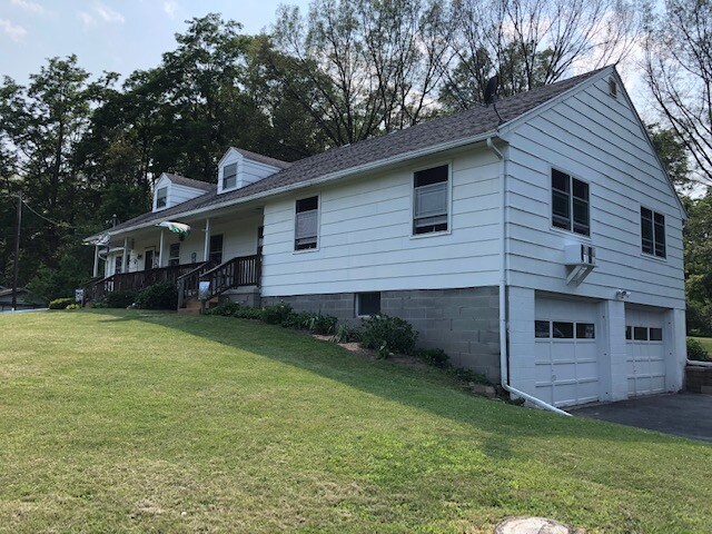 A charming one-story house is set on a gentle hillside, featuring a parking area and a garage. A wide front porch is supported by wooden railings, and large windows provide natural light. Green grass surrounds the home, complementing the tranquil country setting.