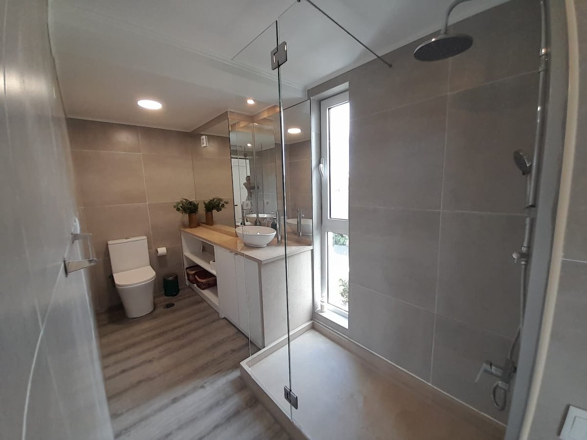 A modern bathroom features a spacious walk-in shower with a clear glass partition. A minimalist vanity with a vessel sink is surrounded by natural light from a nearby window. Neutral grey tiles adorn the walls and floor, contributing to a clean and serene environment.