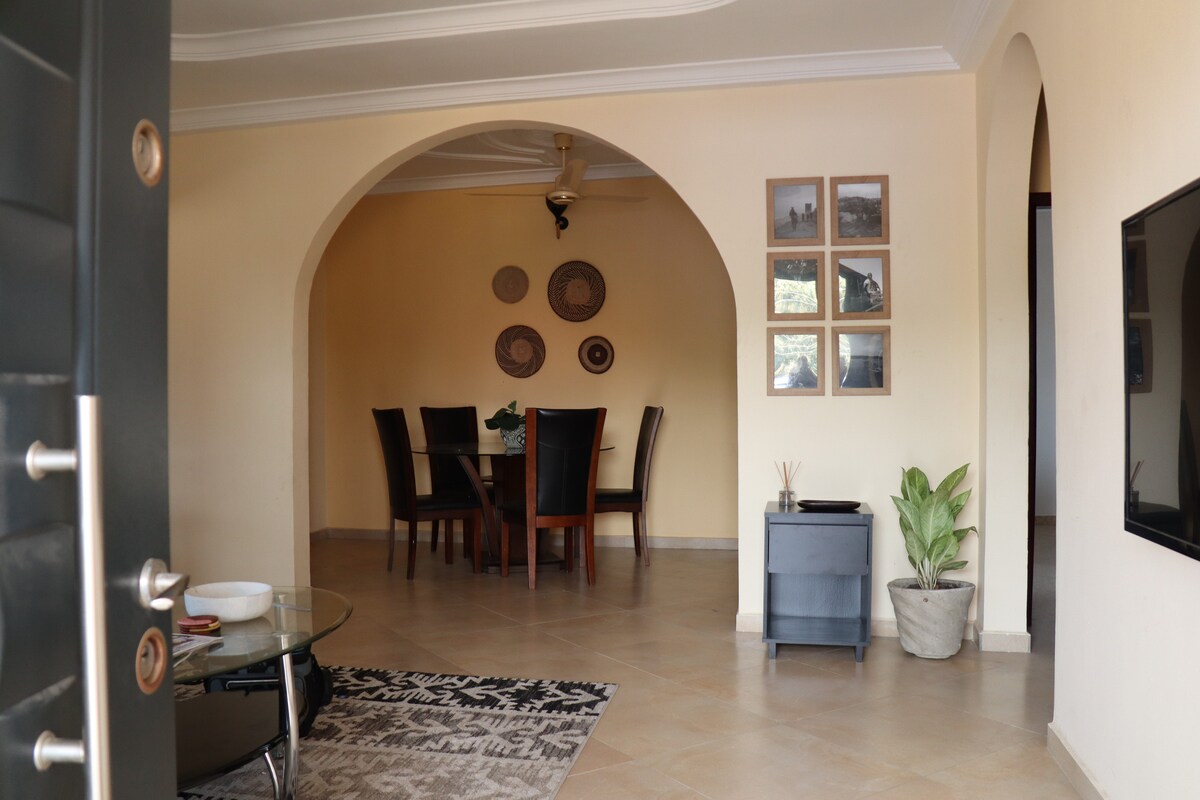 A welcoming entrance reveals an open living area, featuring an arched doorway. A dining table with six chairs is positioned under light fixtures, surrounded by decorative wall art and woven baskets. A potted plant adds a touch of greenery next to a minimalist storage unit.