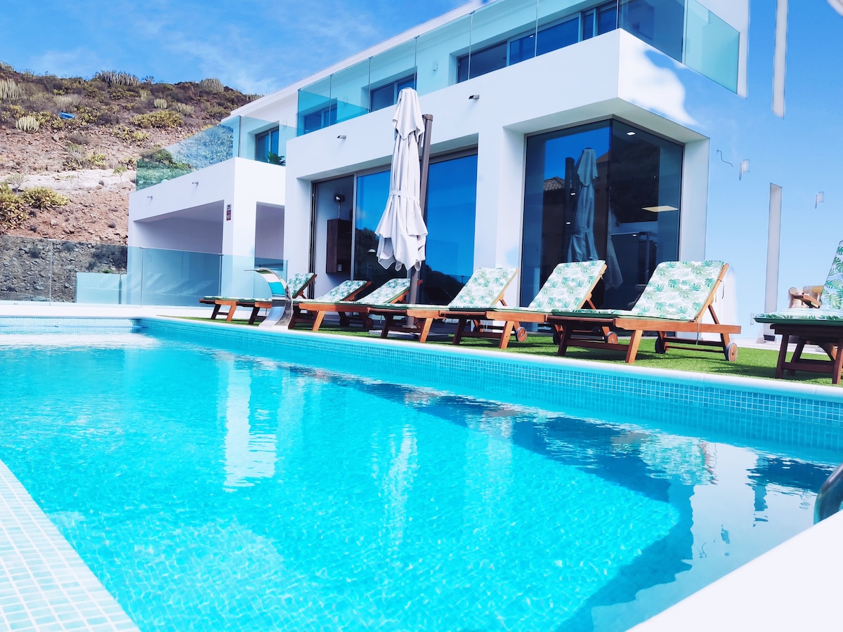 A modern villa features a tranquil heated pool with crystal-clear water. Sun loungers are arranged neatly beside the pool, complemented by a large outdoor umbrella. Floor-to-ceiling windows create an inviting atmosphere, showcasing the scenic surroundings.