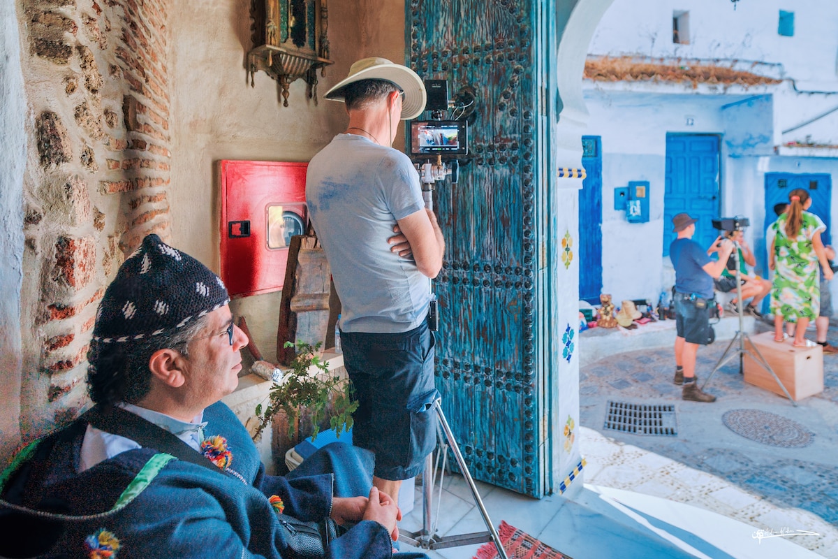 A doorway is highlighted, showing a scene of individuals involved in filming. Outside, vibrant blue walls and traditional architecture are visible, while a seated figure in traditional attire observes the activity. The ground is paved with stones, adding to the cultural ambiance.