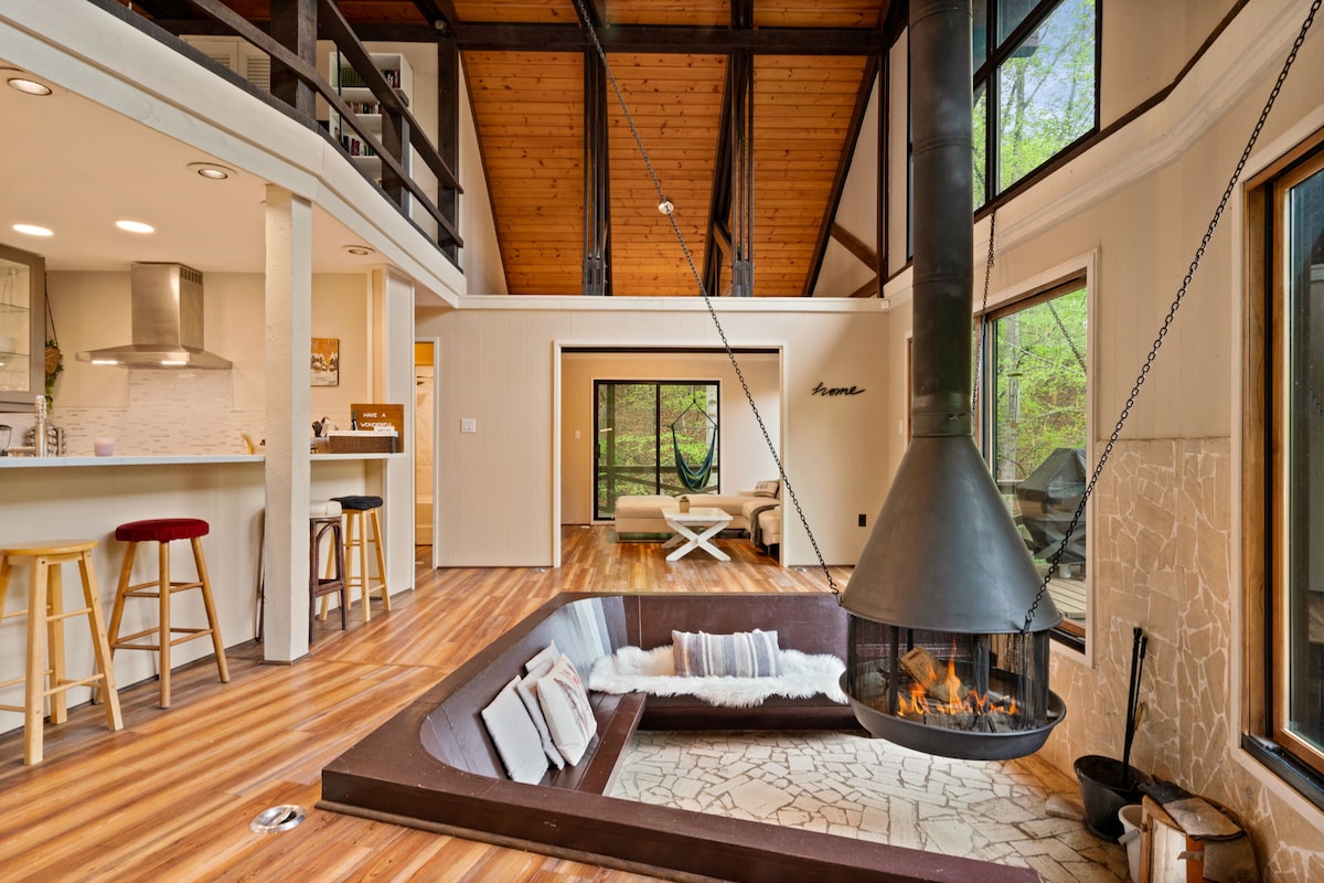 A spacious living area is highlighted by vaulted cathedral ceilings and large windows offering views of the surrounding nature. A unique sunken conversation pit with comfortable cushions surrounds a floating fireplace, creating a cozy centerpiece for gatherings. The open layout seamlessly connects to the kitchen.