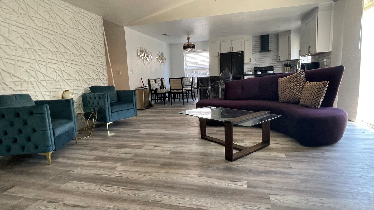 The open living area features contemporary furnishings, including a large purple sectional sofa and two teal armchairs. A glass coffee table sits in the center on a light-colored wooden floor, with the dining area visible in the background, showcasing a table surrounded by six chairs.