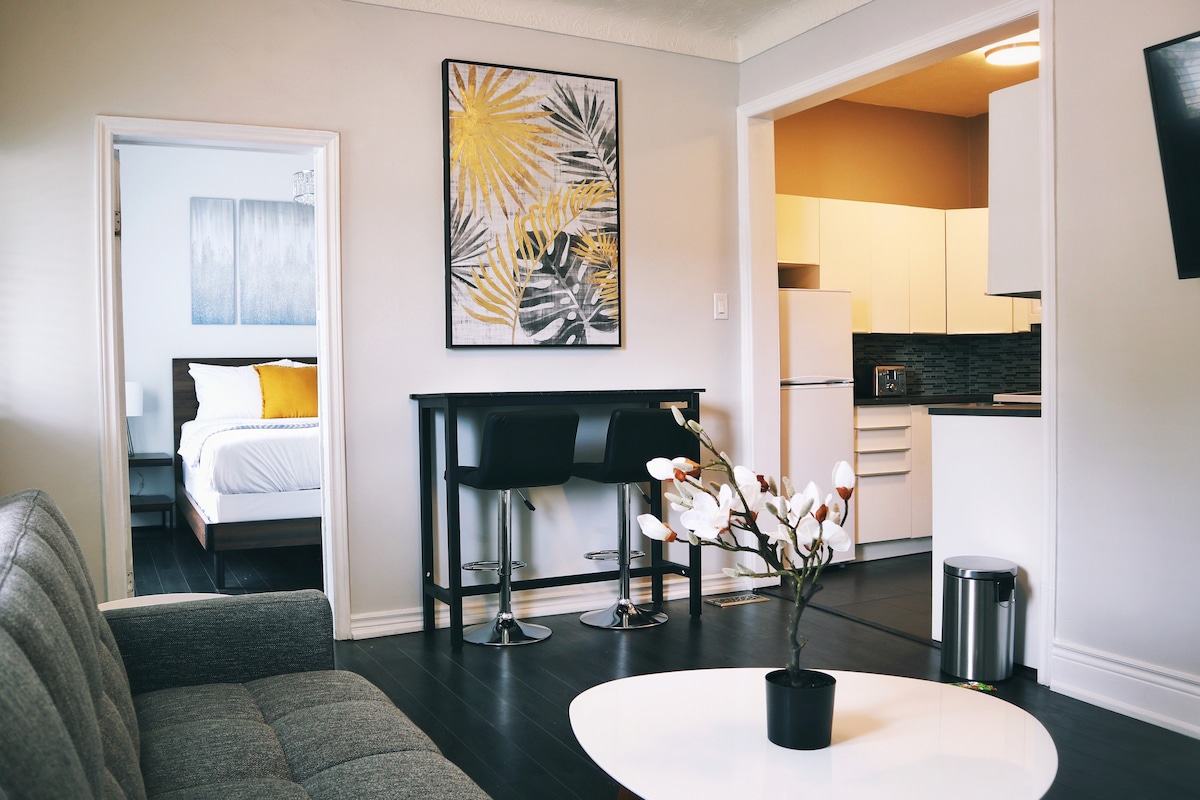 An open living area is showcased, featuring a comfortable gray sofa and a round white coffee table. A small dining table with two black stools is positioned near the sleek kitchen, which includes modern appliances. Bright wall art adds a touch of color to the neutral decor.