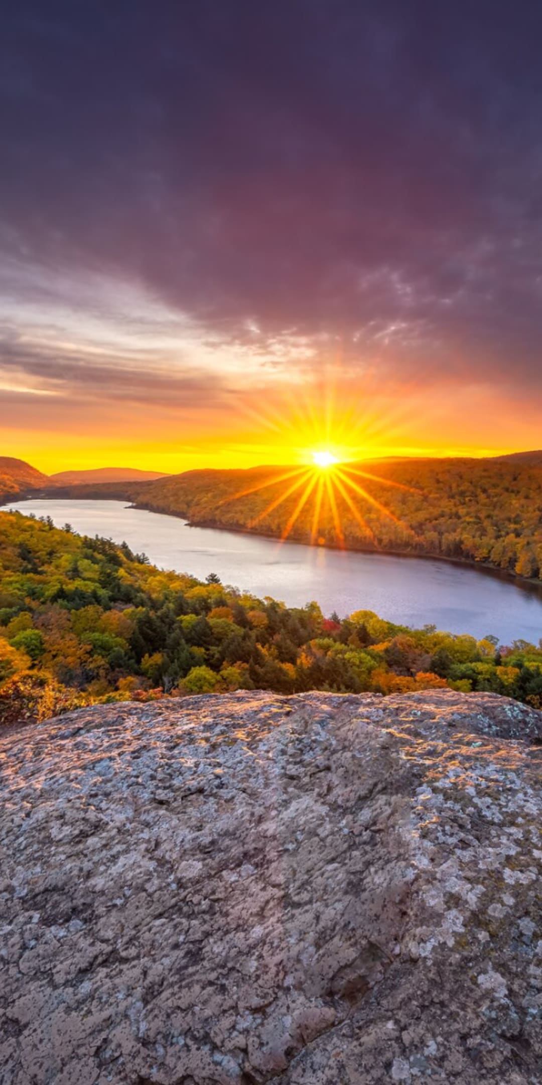 A breathtaking vista is captured, showcasing a river winding through lush greenery, illuminated by a vibrant sunset. The horizon is bathed in warm shades of orange and purple, with rugged rocks in the foreground adding depth to the serene landscape.