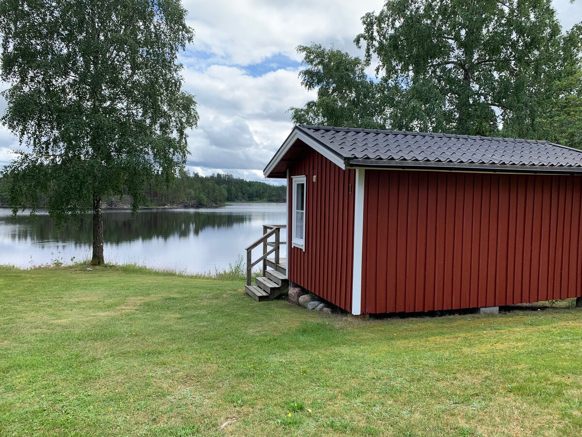 A charming red cabin is positioned beside a serene lake, surrounded by lush green grass and trees. Steps lead from the cabin entrance down to the water's edge, offering a peaceful view of the calm lake surface and the sky reflected on it.