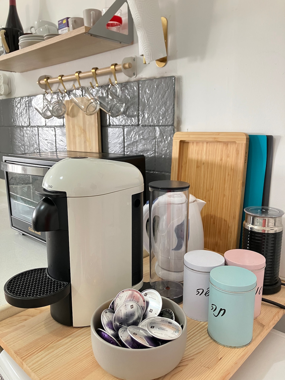 A kitchen countertop displays a coffee machine alongside various coffee pods in a round bowl. A kettle and a glass container are positioned nearby. Wall shelves hold tea mugs and additional kitchen items, offering a functional and organized space.